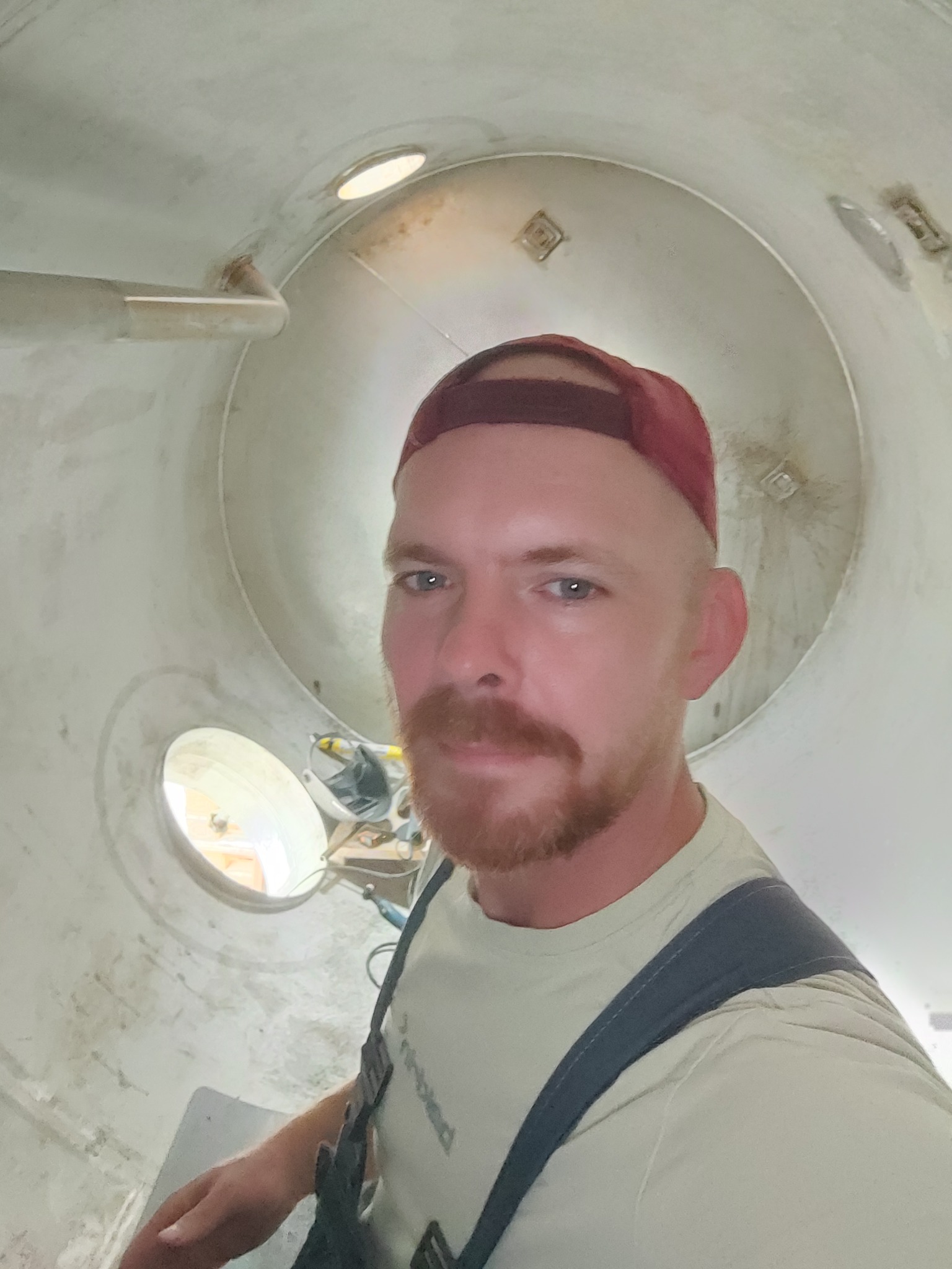 Spawacz wewnątrz metalowej rury, widoczne narzędzia i otwór oświetleniowy,  w burgundowej czapce z daszkiem,  spogląda w obiektyw.