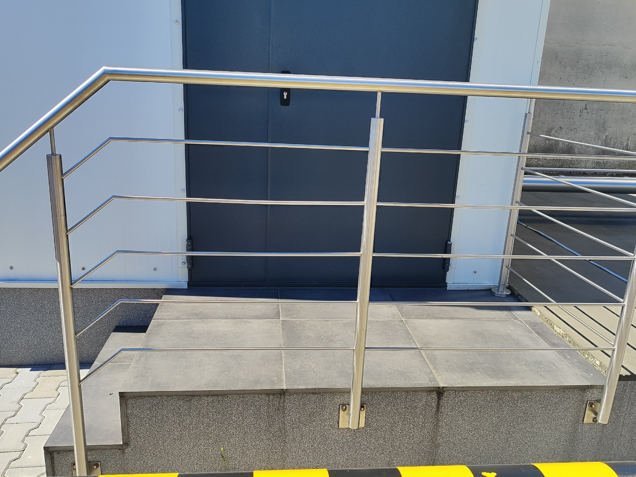 Nowoczesna, metalowa poręcz schodowa ze stali nierdzewnej, z poziomymi prętami, zamontowana przy wejściu do budynku z szarymi drzwiami i schodami wyłożonymi płytkami.