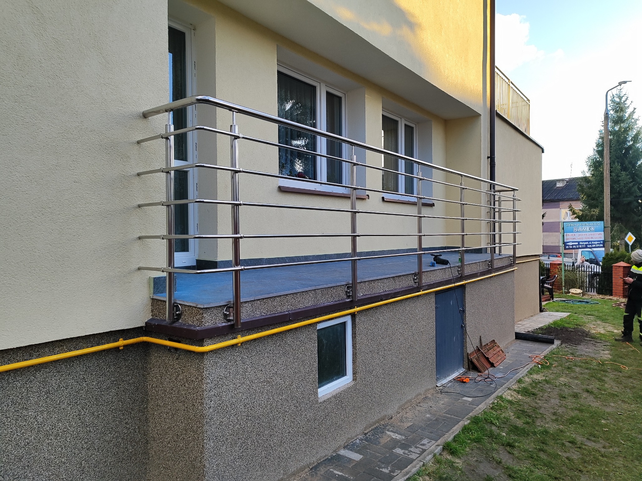 Balkon z balustradą ze stali nierdzewnej, widok z zewnątrz. Jasna elewacja budynku, widoczne okna i fragment instalacji gazowej. Ujęcie z dołu, w tle zieleń.