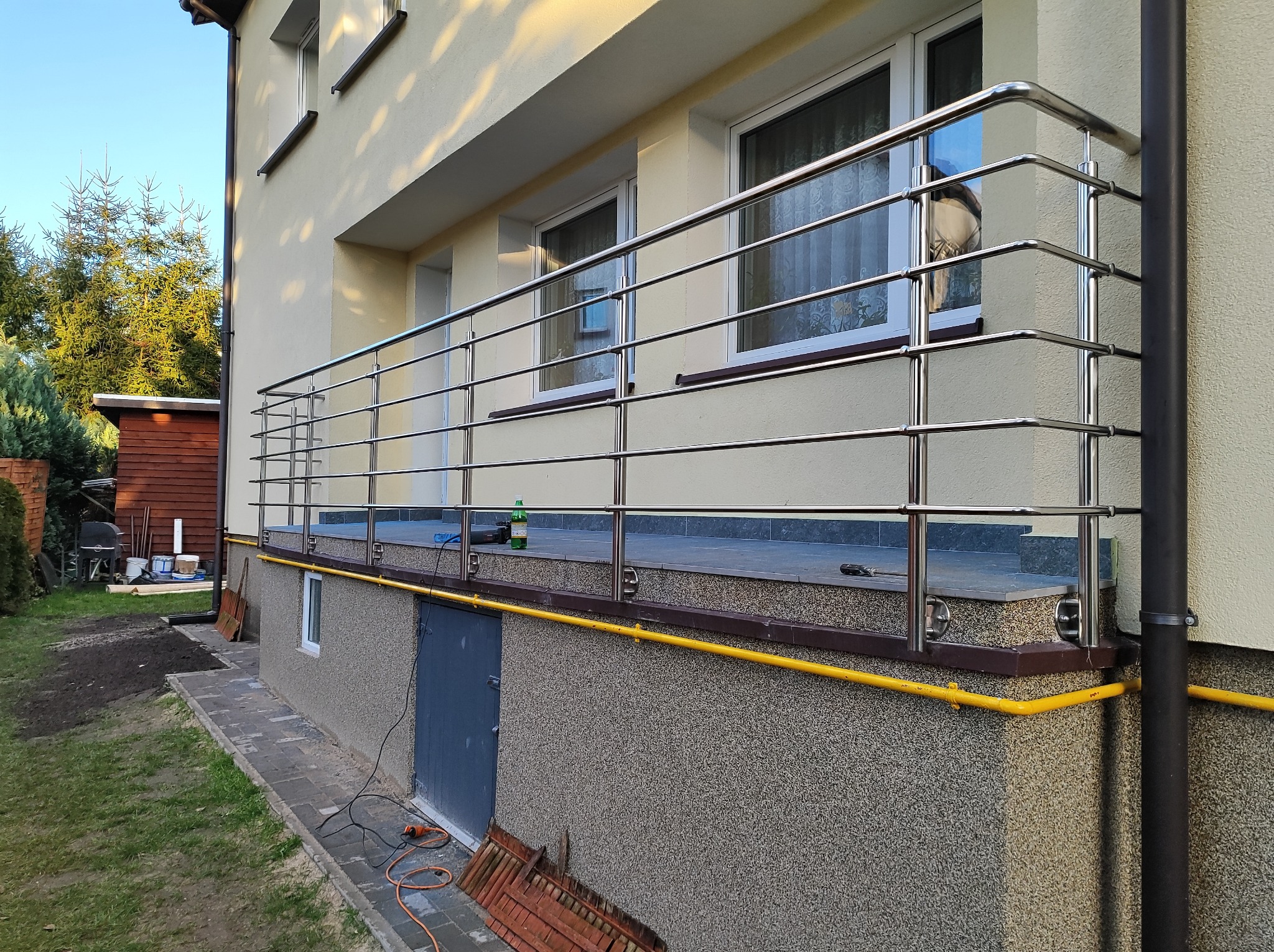 Balkon z balustradą ze stali nierdzewnej, widok z dołu. Na balkonie narzędzia i butelka. Żółta rura gazowa wzdłuż ściany. Trawa i drzewa w tle.