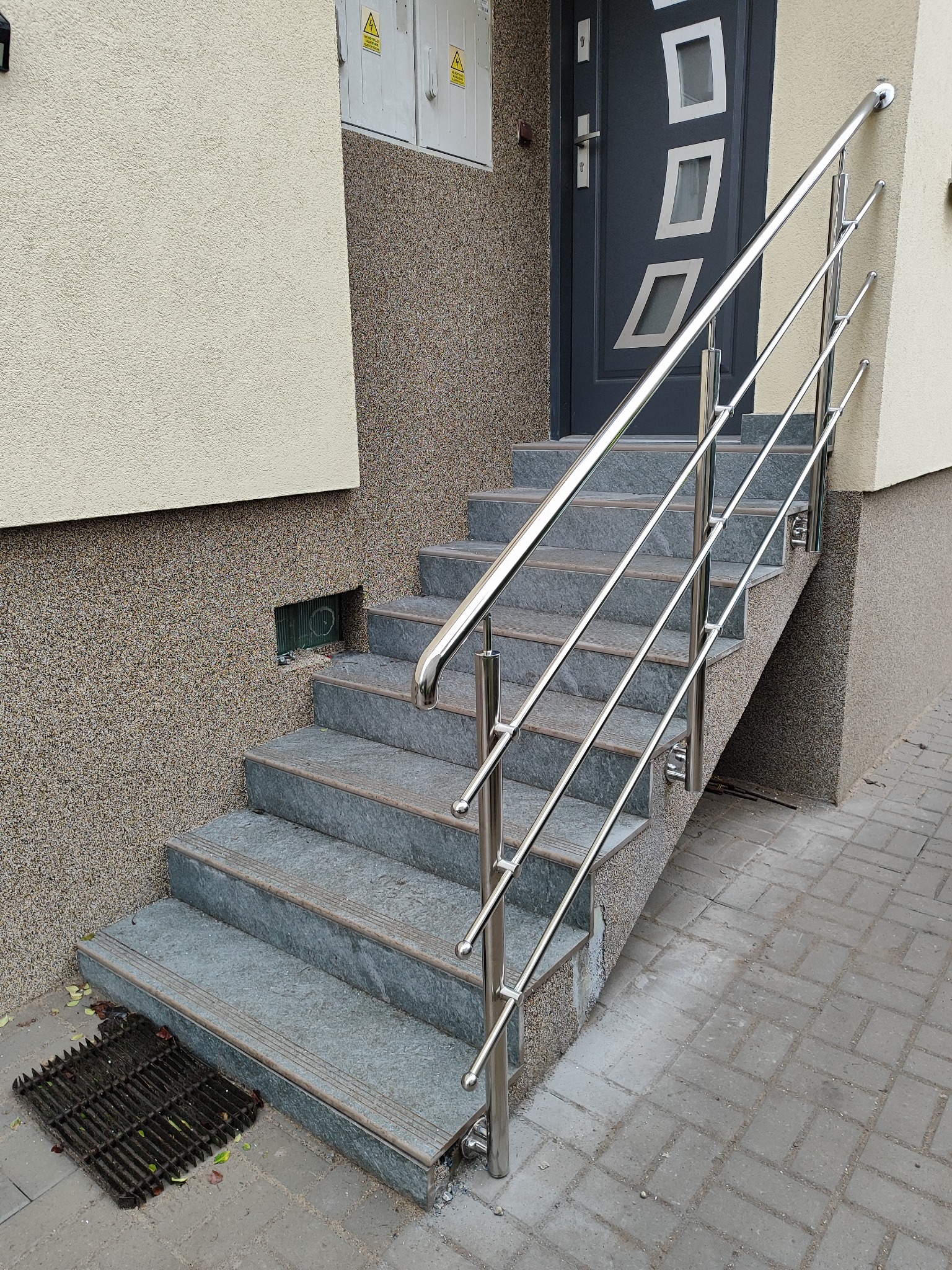 Schody wejściowe z szarych płytek z nowoczesną, chromowaną poręczą. Widoczne drzwi wejściowe z geometrycznymi zdobieniami i fragment chodnika z kostki brukowej.