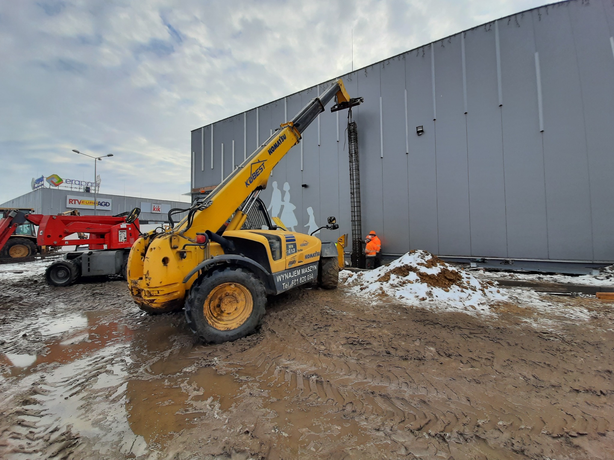 Żółty teleskopowy wózek widłowy Komatsu Kobest podnosi zbrojenie na tle szarej ściany budynku, widoczny pracownik w pomarańczowej kamizelce, teren budowy z błotem i śniegiem.