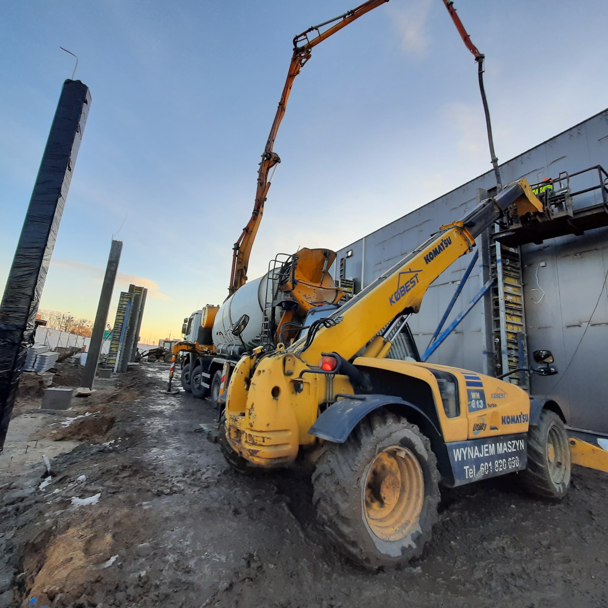 Żółta ładowarka teleskopowa Komatsu z napisem 'WYNAJEM MASZYN' na tle betonomieszarki z wysięgnikiem podczas wylewania betonu na placu budowy z przygotowanymi słupami.