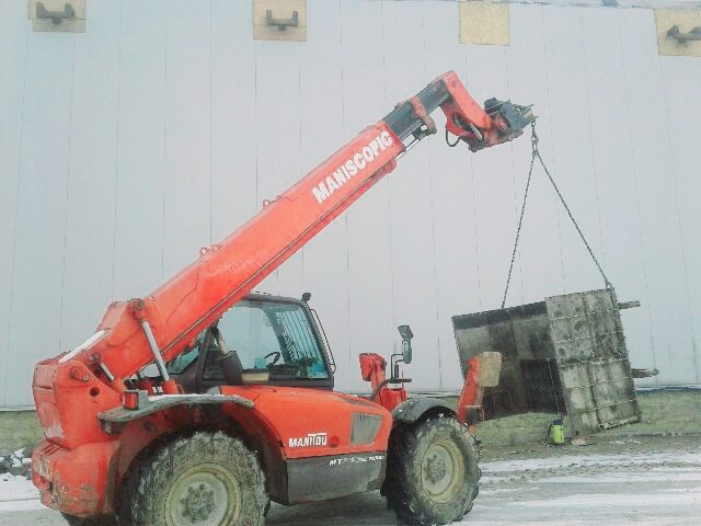 Czerwona ładowarka teleskopowa Manitou podnosi formę szalunkową na tle szarej ściany magazynu.