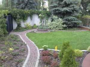 Zadbany ogród z równo przystrzyżonym trawnikiem, ścieżką z czerwonej kostki brukowej, ozdobnymi krzewami i wysokimi iglakami na tle białego muru.