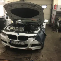 Białe BMW z otwartą maską w warsztacie; widoczny silnik, butla z gazem i narzędzia w tle.