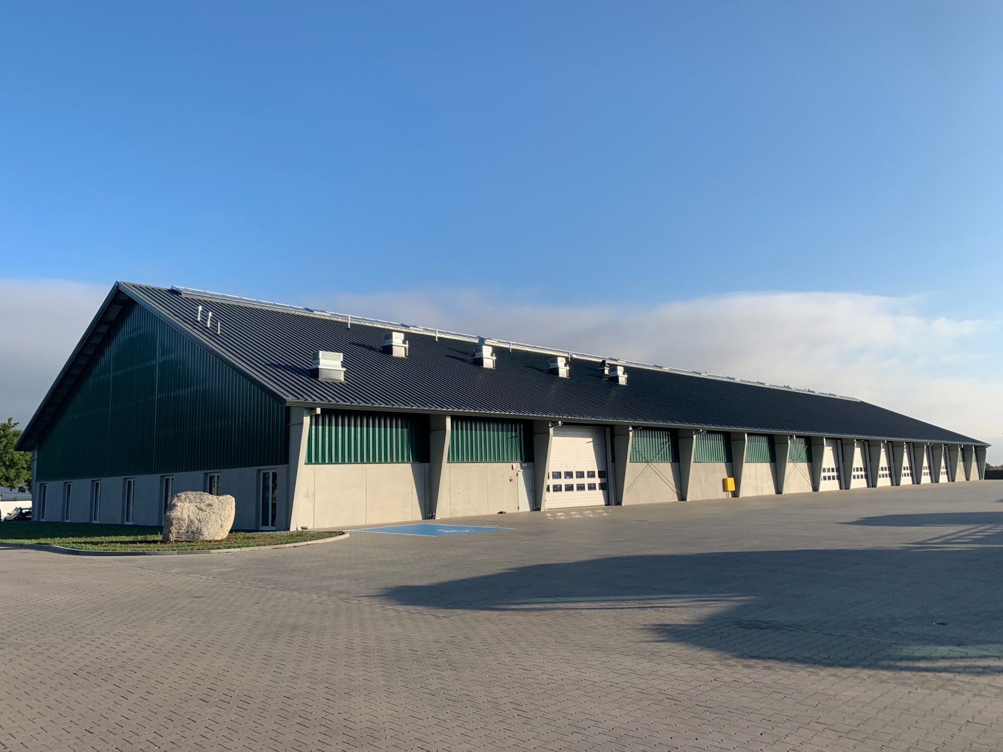 Długi, parterowy budynek magazynowy z licznymi bramami wjazdowymi, zielonymi panelami i ciemnym dachem, widziany w słoneczny dzień.