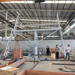 HM-SOLAR - Wnętrze hali produkcyjnej z suwnicą podnoszącą metalową konstrukcję wsporczą. Trzy osoby obserwują proces, a w tle widoczne są elementy konstrukcyjne i skrzynie z falownikami PV Growatt.