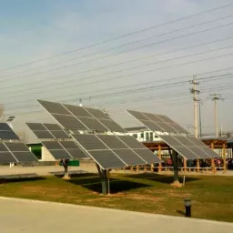 Rząd paneli słonecznych ustawionych na trawniku, wspartych na pojedynczych metalowych słupach, widoczne linie wysokiego napięcia na tle pochmurnego nieba.