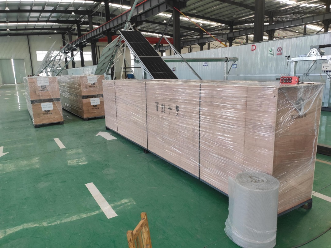 Palety z panelami słonecznymi, zabezpieczone folią, w hali produkcyjnej z zieloną podłogą i białymi pasami, w tle widoczny system montażowy paneli.