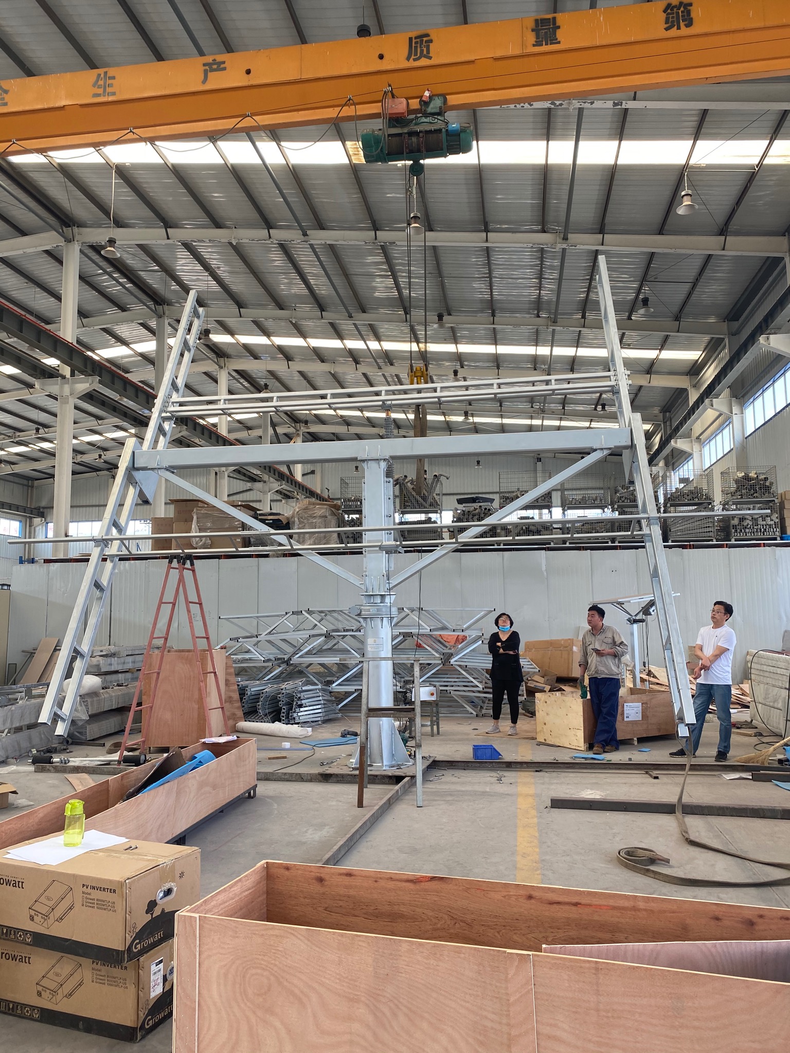 Wnętrze hali produkcyjnej z suwnicą podnoszącą metalową konstrukcję wsporczą. Trzy osoby obserwują proces, a w tle widoczne są elementy konstrukcyjne i skrzynie z falownikami PV Growatt.