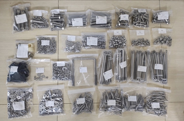 Zestaw elementów montażowych w przezroczystych woreczkach, zawierający śruby, nakrętki, podkładki, obejmy U-kształtne i inne drobne części, ułożonych na jasnej powierzchni.