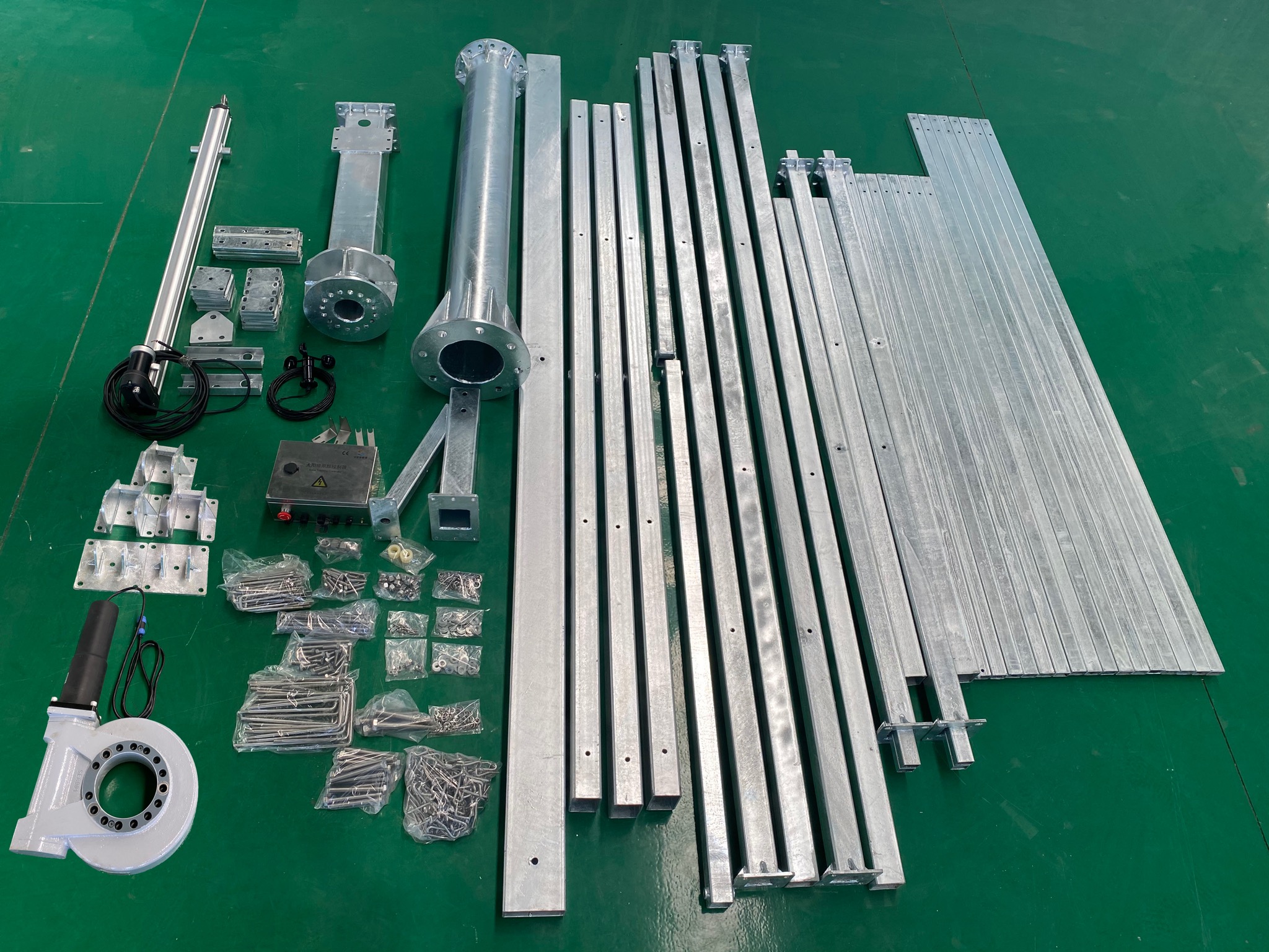 Elementy konstrukcyjne stalowe, w tym rury, profile, śruby i złączki, rozłożone na zielonym tle, prawdopodobnie przygotowane do montażu systemu.