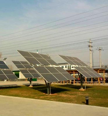 Rząd paneli słonecznych ustawionych na trawniku, wspartych na pojedynczych metalowych słupach, widoczne linie wysokiego napięcia na tle pochmurnego nieba.