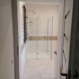 Kajdos Mariusz - Nowoczesna łazienka z kabiną prysznicową typu walk-in, widok z korytarza przez otwarte drzwi, białe płytki z poziomym paskiem dekoracyjnym, grzejnik drabinkowy w kolorze antracytowym.