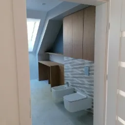 Nowoczesna łazienka na poddaszu z białą ceramiką, drewnianymi szafkami i biurkiem, oraz wzorzystymi płytkami na ścianie, widziana zza uchylonych drzwi.