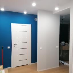 Kajdos Mariusz - Świeżo pomalowane wnętrze mieszkania z białą stolarką drzwiową i intensywnie niebieską ścianą, widoczne ślady prac malarskich przy podłodze i narzędzia malarskie w tle.