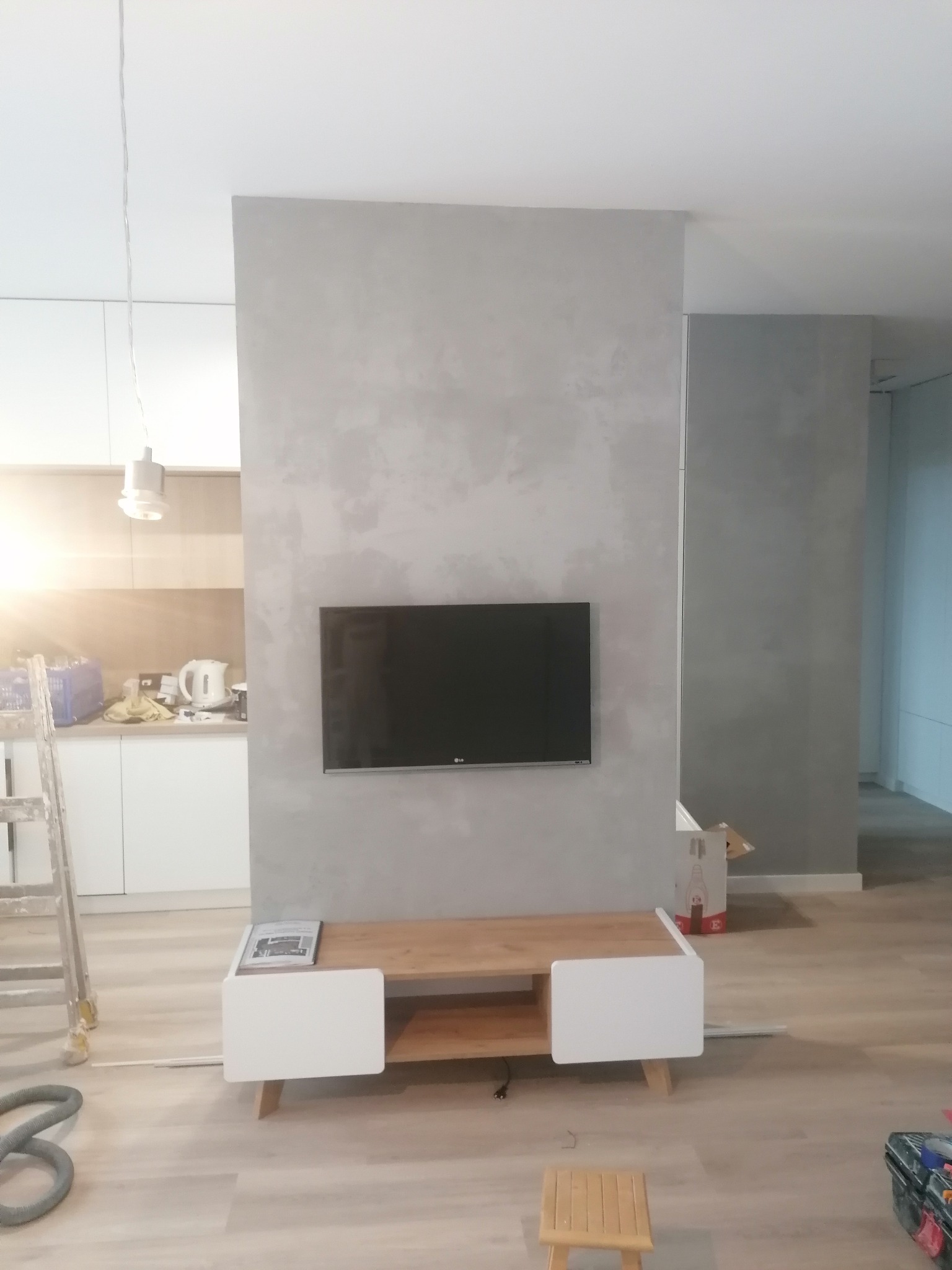 Ściana w salonie po nałożeniu dekoracyjnej szarej gładzi, z widocznym telewizorem i stolikiem RTV, drabina malarska oparta o ścianę, resztki materiałów budowlanych na podłodze, lampa wisząca.