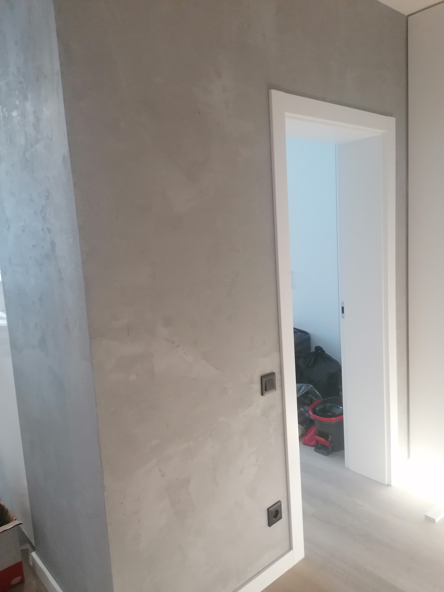 Ściana z szarą dekoracyjną strukturą, widoczne dwa gniazdka elektryczne, biała ościeżnica drzwi, w tle wiadro i worki na śmieci.