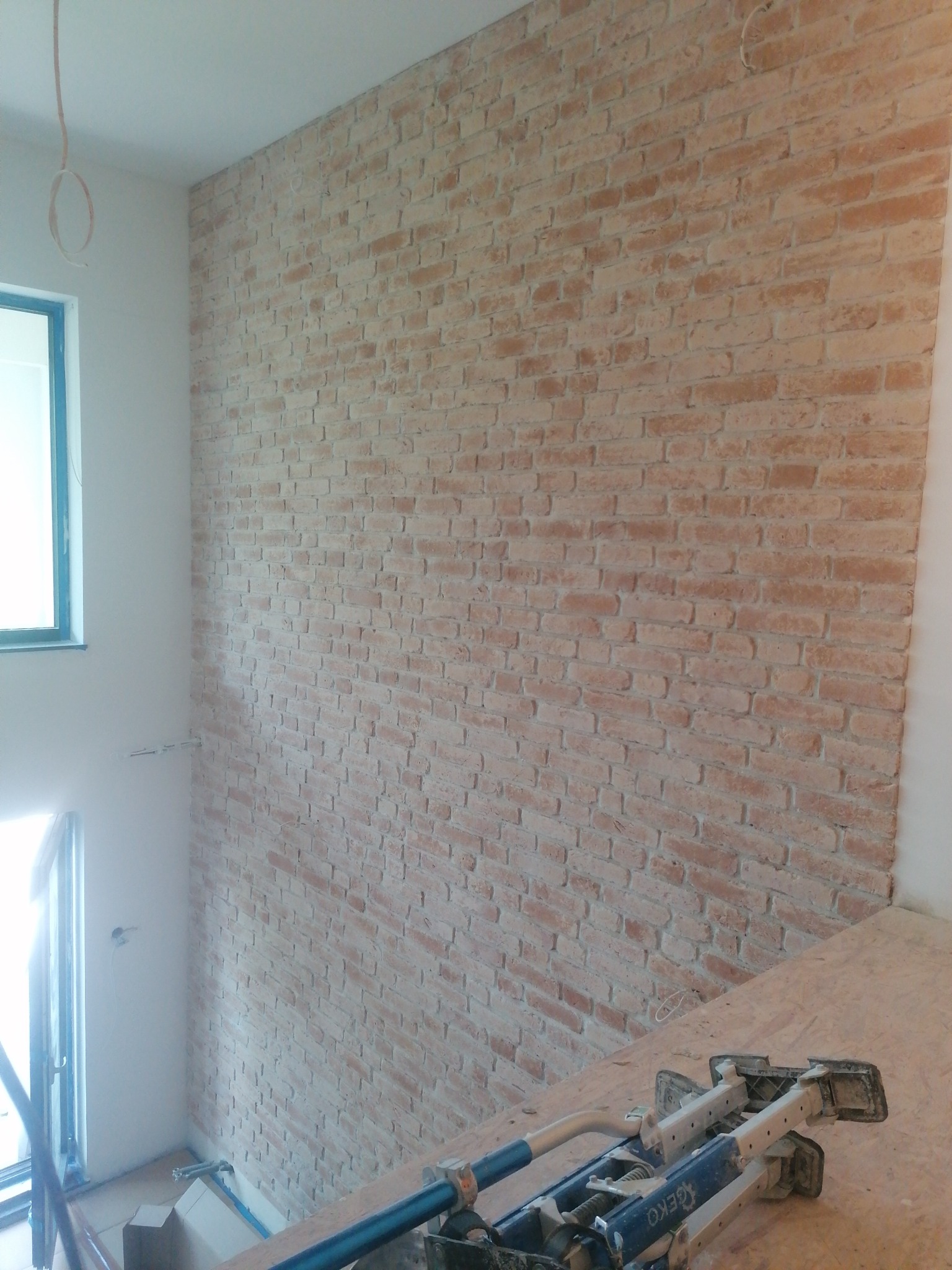 Ściana z cegły dekoracyjnej w jasnym odcieniu, widok z góry z rusztowaniem na pierwszym planie, surowy kabel zwisający z sufitu.