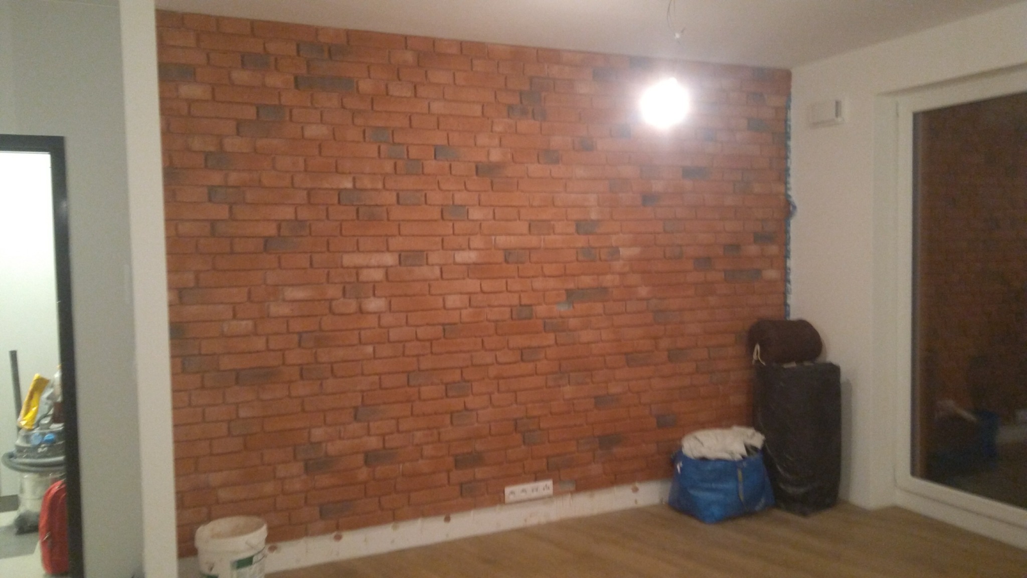 Ściana wykończona cegłą dekoracyjną w salonie, widoczne gniazdka elektryczne przy podłodze, worki budowlane i sprzęt czyszczący w tle.
