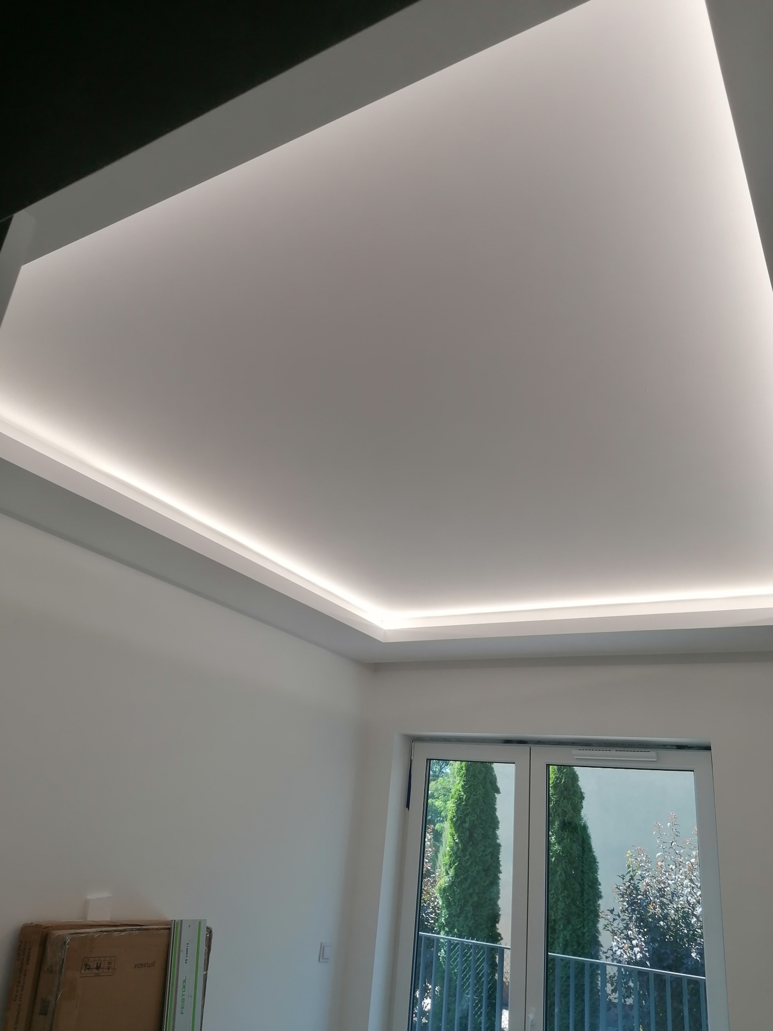 Nowoczesny sufit podwieszany z oświetleniem LED, widok z dołu, w tle okno z widokiem na zieleń.