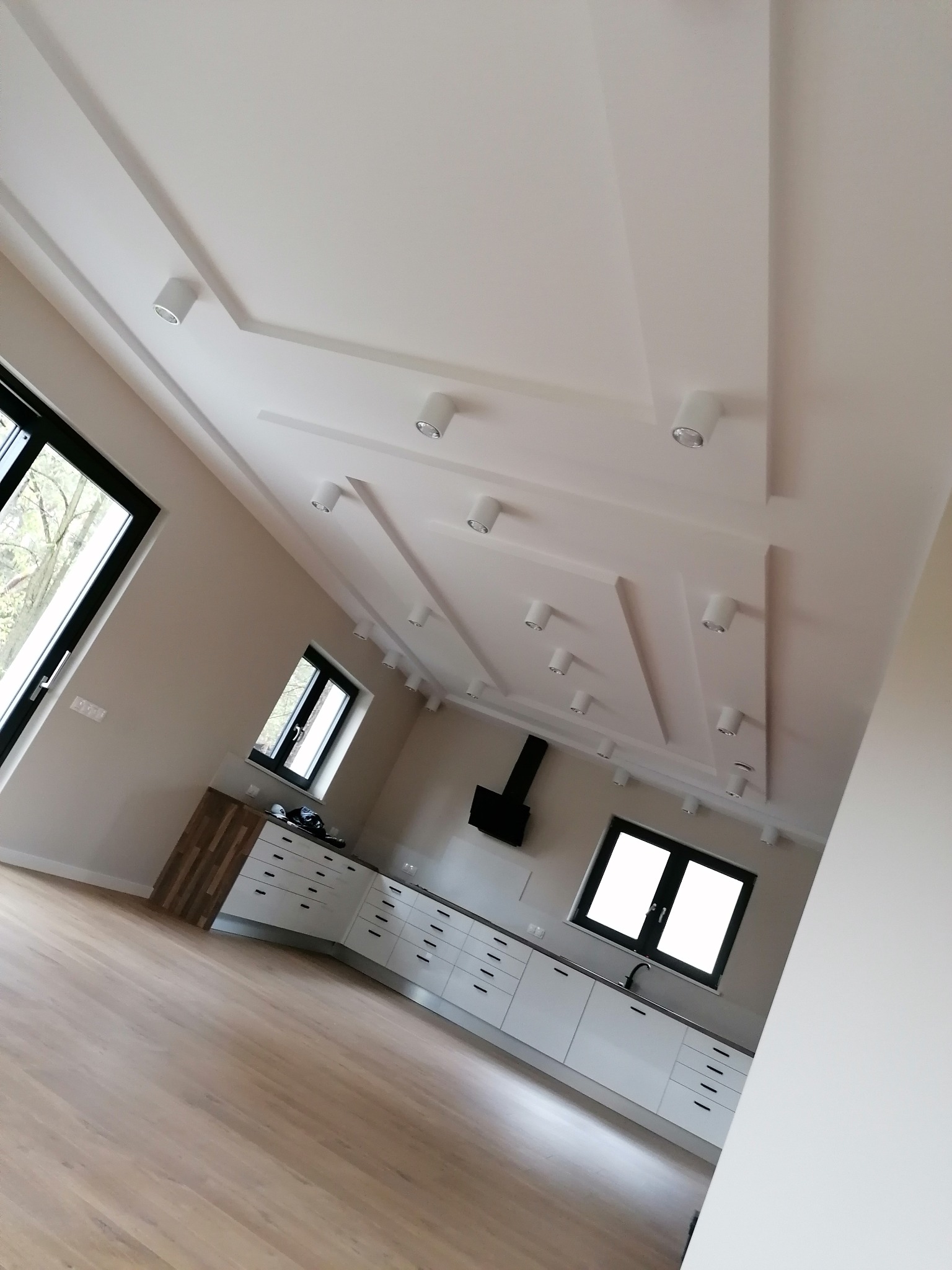 Nowoczesna kuchnia w jasnych barwach z drewnianą podłogą, białymi szafkami i oryginalnym sufitem z oświetleniem punktowym.