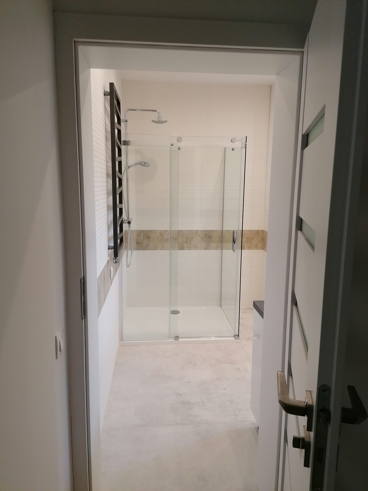 Nowoczesna łazienka z kabiną prysznicową typu walk-in, widok z korytarza przez otwarte drzwi, białe płytki z poziomym paskiem dekoracyjnym, grzejnik drabinkowy w kolorze antracytowym.