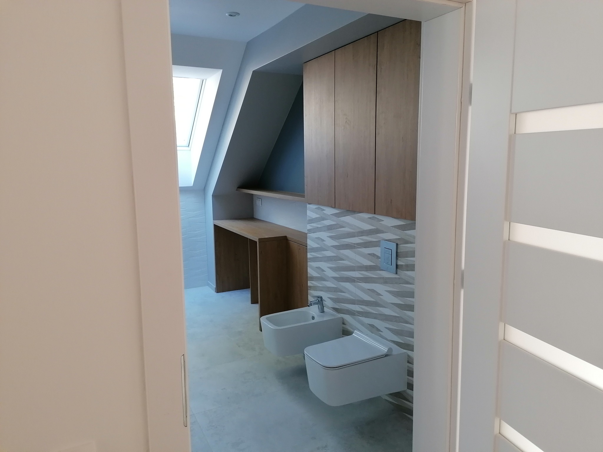 Nowoczesna łazienka na poddaszu z białą ceramiką, drewnianymi szafkami i biurkiem, oraz wzorzystymi płytkami na ścianie, widziana zza uchylonych drzwi.