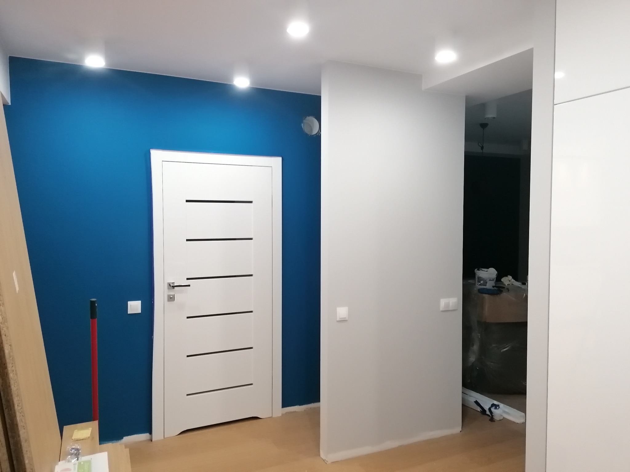 Świeżo pomalowane wnętrze mieszkania z białą stolarką drzwiową i intensywnie niebieską ścianą, widoczne ślady prac malarskich przy podłodze i narzędzia malarskie w tle.