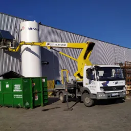 Żółty podnośnik koszowy na tle szarej, blaszanej elewacji przemysłowej; platforma robocza z pracownikiem przy białym zbiorniku Messer; obok zielony kontener na odpady Lubelskiej Agencji Ochrony...