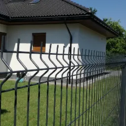 Ciemnoszary panelowy płot z widocznymi słupkami na tle białego domu z brązowymi okiennicami i zielonym trawnikiem.