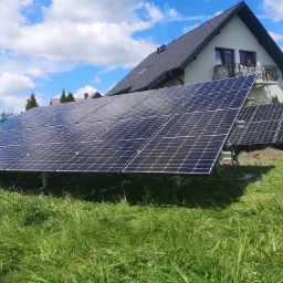 Panele słoneczne zamontowane na trawiastym terenie w Skoczowie, z widocznym domem jednorodzinnym w tle oraz błękitnym niebem z chmurami.