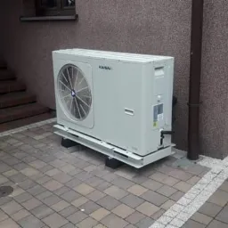 Zewnętrzna jednostka pompy ciepła marki Kaisai, zamontowana na metalowej konstrukcji na kostce brukowej, widoczna ściana budynku z brązowym tynkiem strukturalnym i schody wejściowe.