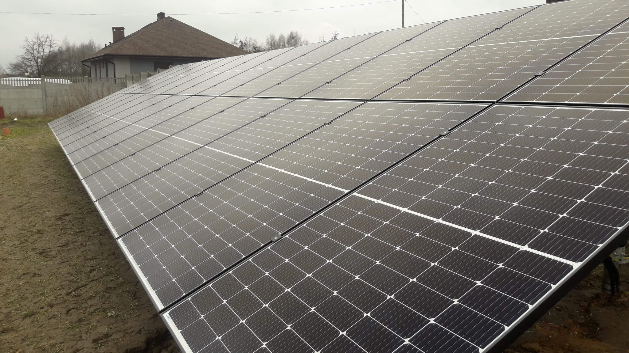 Instalacja paneli słonecznych na gruncie obok domu jednorodzinnego z brązowym dachem, pochmurne niebo w tle.