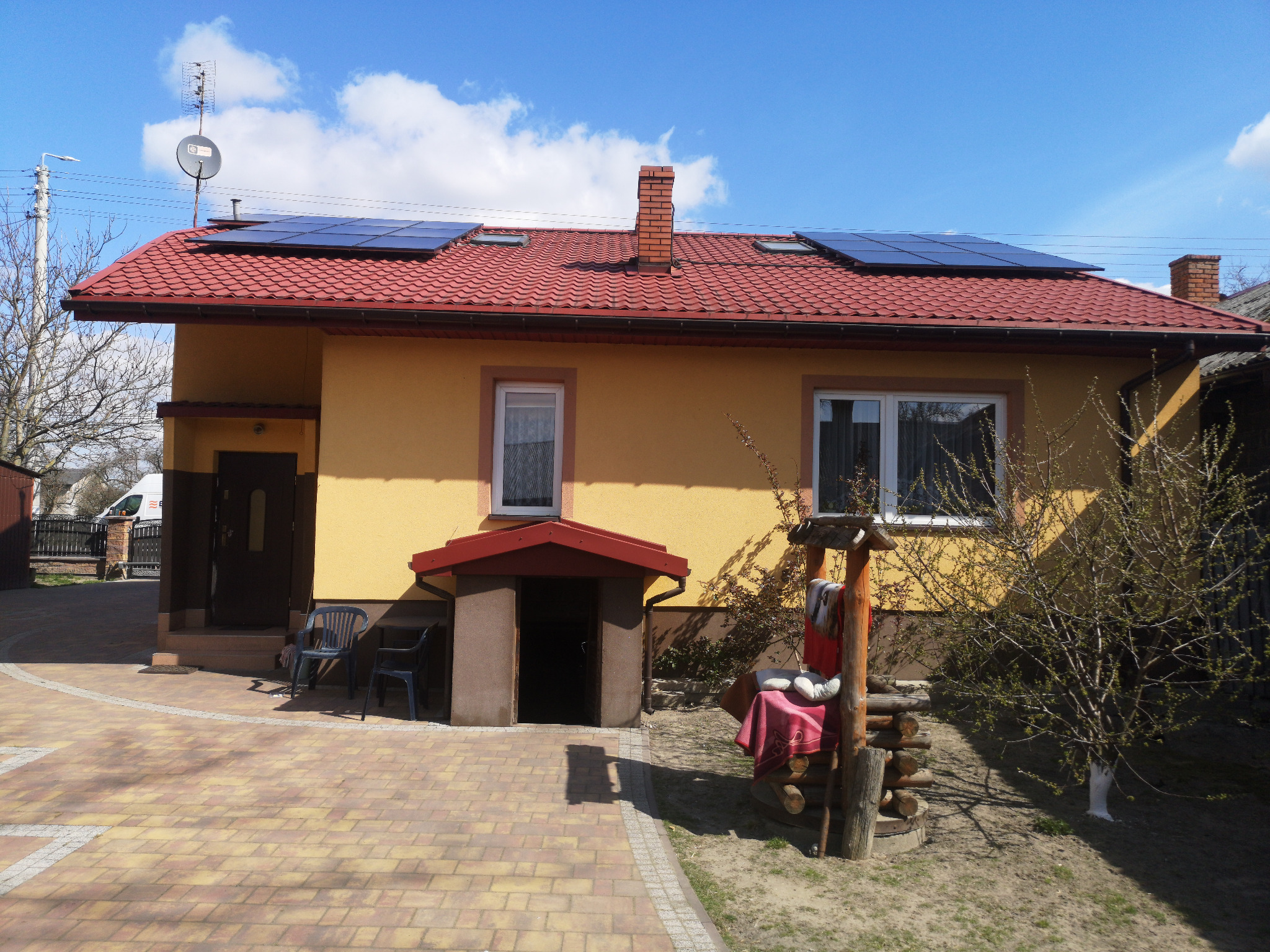 Żółty dom jednorodzinny z czerwoną dachówką, na której zamontowane są panele fotowoltaiczne; na dachu widoczna antena satelitarna oraz komin; na podwórku studnia z drewnianą obudową.