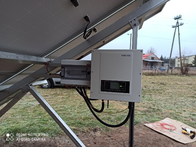 Instalacja paneli słonecznych: widok od spodu na panele, skrzynkę rozdzielczą i falownik SOFAR, z narzędziami i przewodami na kartonie w tle, na zewnątrz budynku.