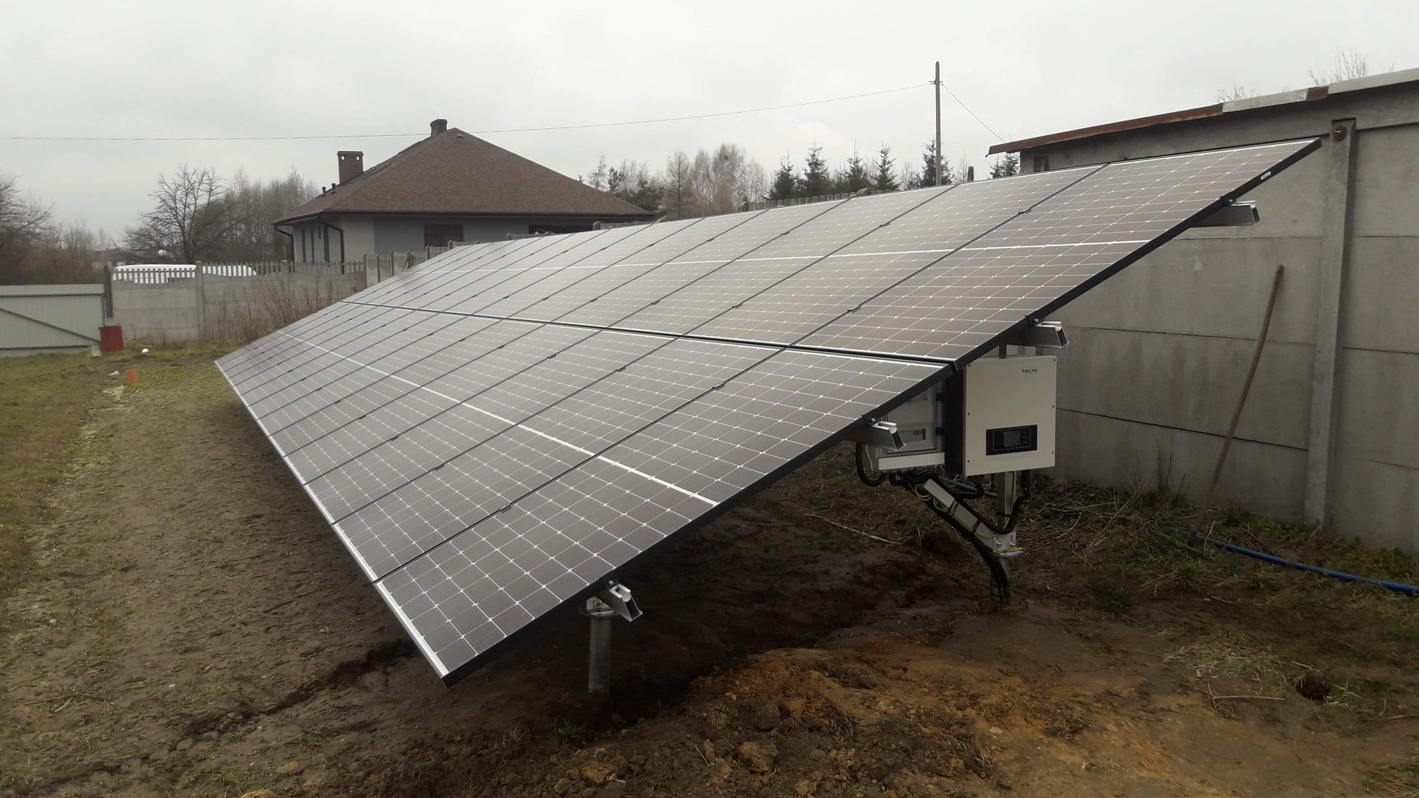 Panel fotowoltaiczny na metalowych podporach, zamontowany na ziemi obok betonowego ogrodzenia i budynku mieszkalnego w pochmurny dzień.