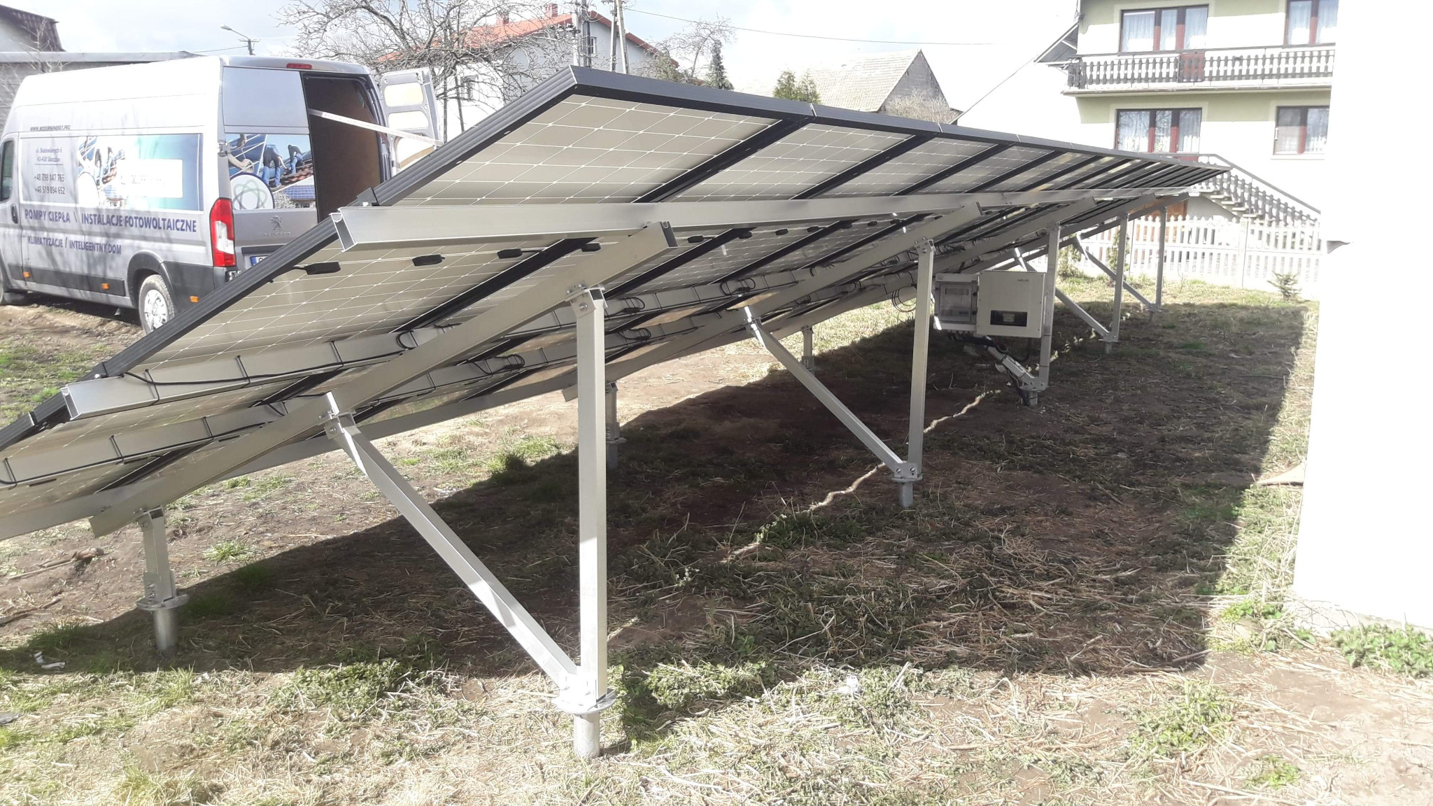 Montaż paneli słonecznych na konstrukcji wsporczej z widocznym inwerterem, na tle budynku mieszkalnego i samochodu dostawczego firmy instalacyjnej.