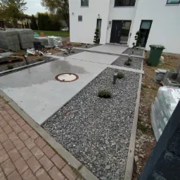 Nowoczesny podjazd do domu z szarych płyt betonowych i strefami wysypanymi szarym żwirem, oświetlony niskimi lampami, widoczne materiały budowlane i zielony kosz na śmieci.