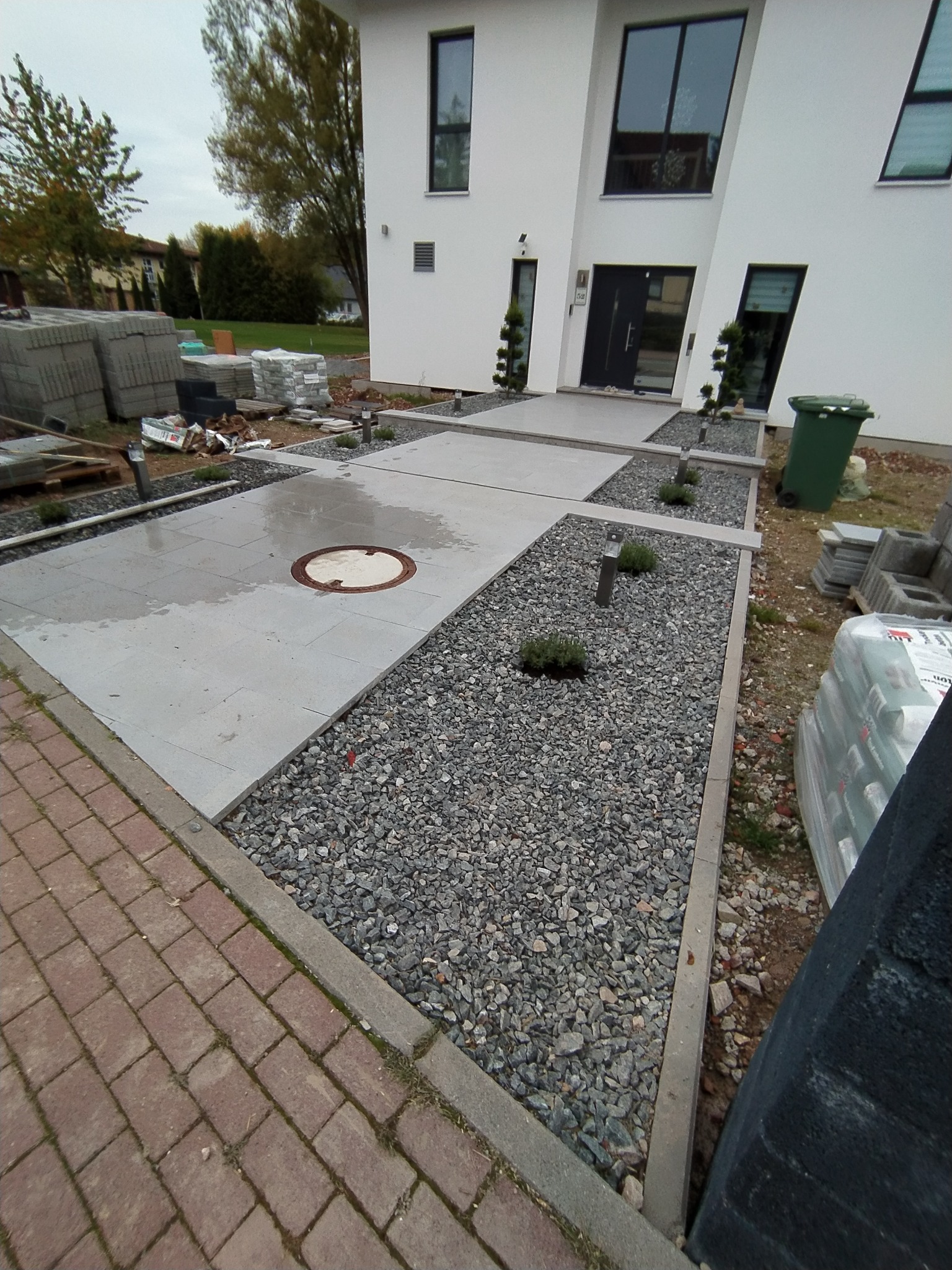 Nowoczesny podjazd do domu z szarych płyt betonowych i strefami wysypanymi szarym żwirem, oświetlony niskimi lampami, widoczne materiały budowlane i zielony kosz na śmieci.