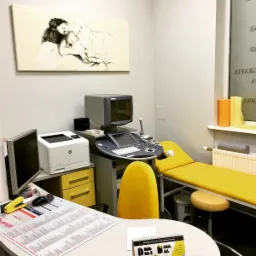 Gabinet lekarski z aparatem USG, żółtym fotelem i leżanką, na ścianie obraz przedstawiający matkę z dzieckiem.