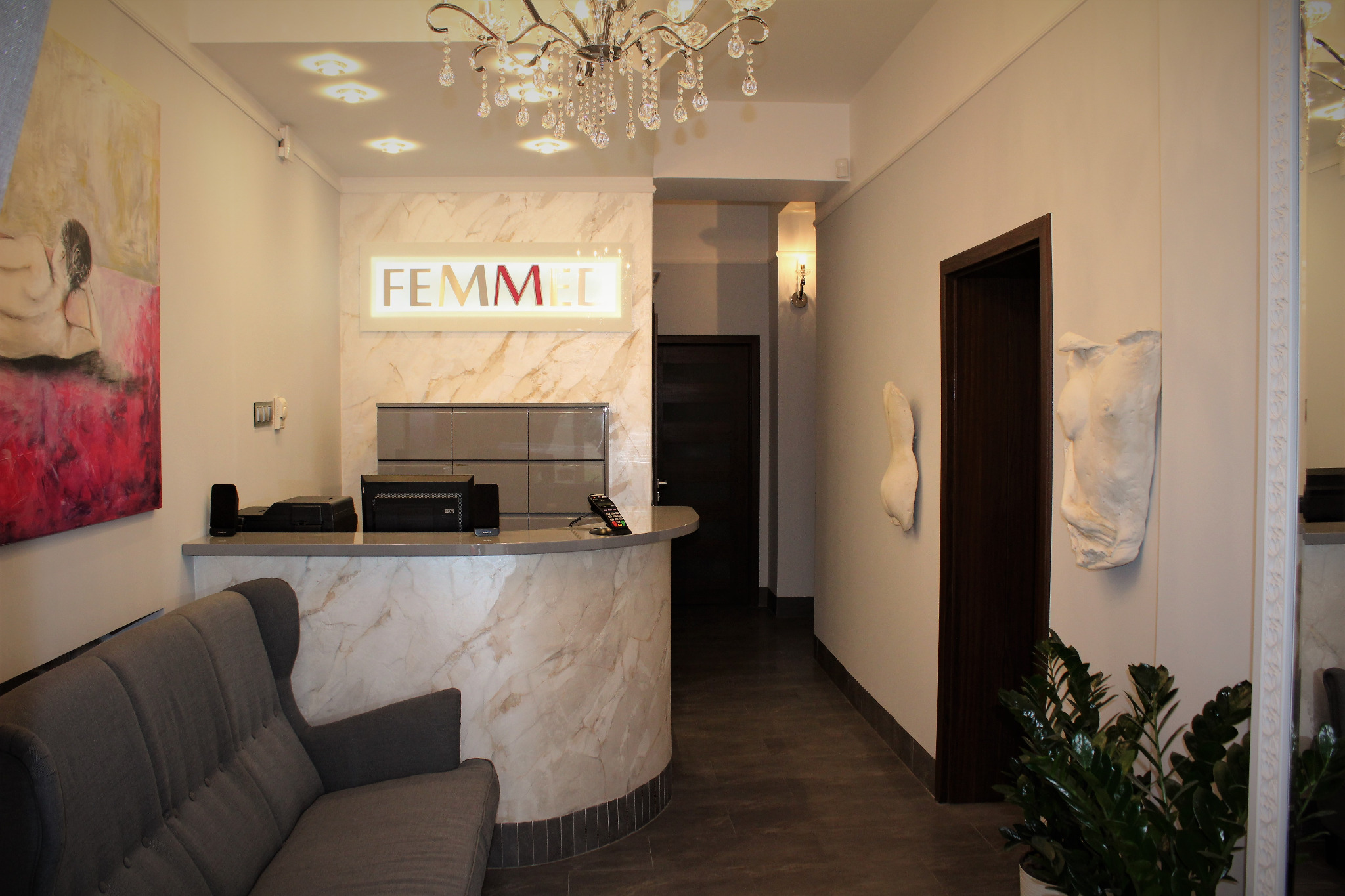Jasna recepcja z marmurowym wykończeniem, szarą sofą i abstrakcyjnym obrazem na ścianie, oświetlona kryształowym żyrandolem.