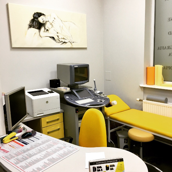 Gabinet lekarski z aparatem USG, żółtym fotelem i leżanką, na ścianie obraz przedstawiający matkę z dzieckiem.