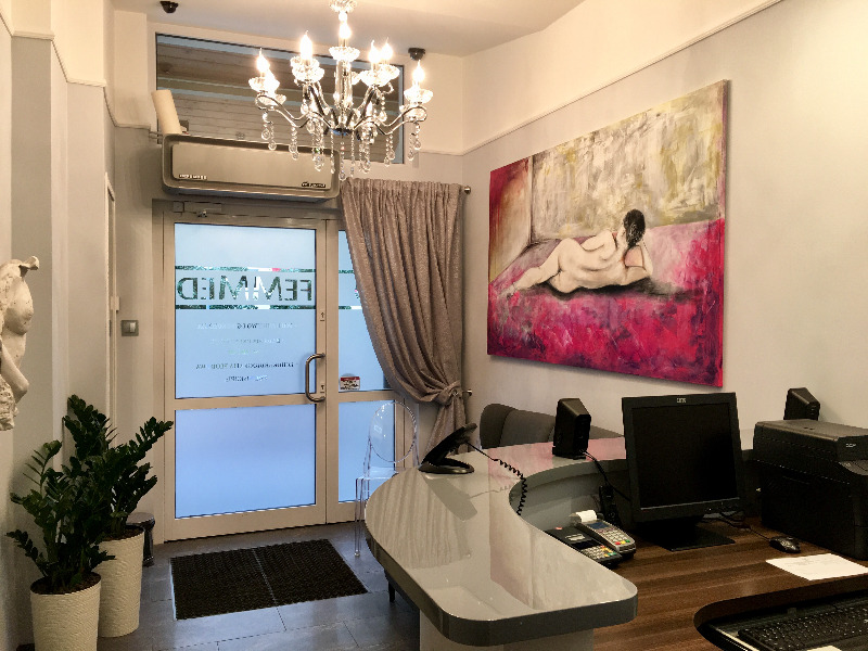 Jasna recepcja z biurkiem o zaokrąglonym kształcie, roślinami w donicach przy wejściu i obrazem aktu na ścianie w tle.