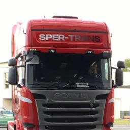 Czerwona ciężar&oacute;wka Scania R450 firmy Sper-Trans z kierowcą w kabinie, widok z przodu na parkingu.