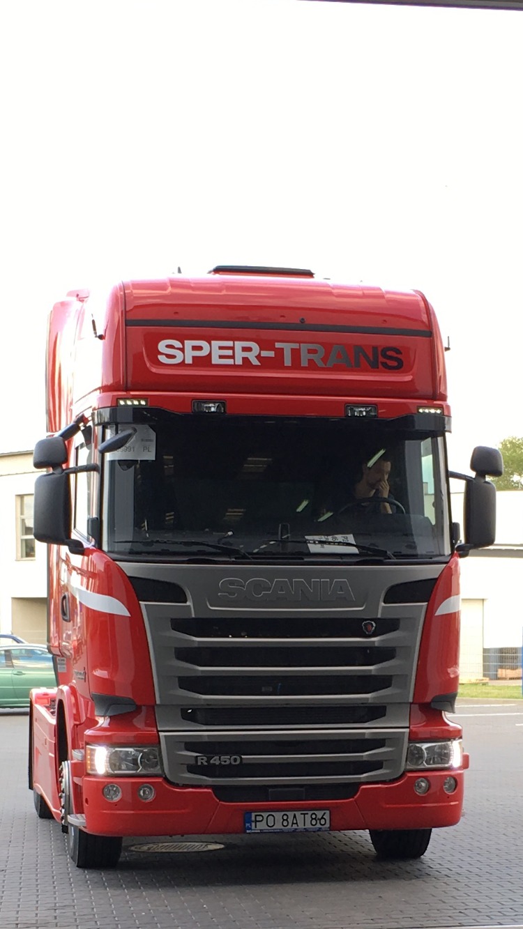 Czerwona ciężarówka Scania R450 firmy Sper-Trans z kierowcą w kabinie, widok z przodu na parkingu.