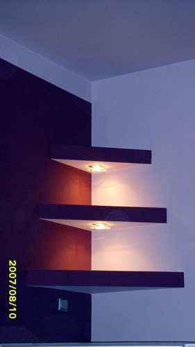 Trzy trójkątne półki narożne z wbudowanym oświetleniem, umieszczone na ścianie pomalowanej w dwóch kolorach: ciemny fiolet i jasny fiolet, w rogu pokoju, widoczne gniazdko elektryczne.