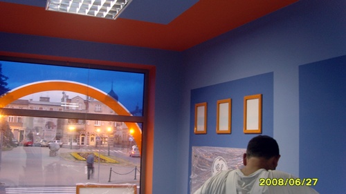 Wnętrze pomalowane na niebiesko z pomarańczowymi akcentami, widoczne trzy puste ramki na ścianie, okno z widokiem na plac miejski i pracownik w białej koszulce podczas remontu.