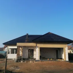 Dom w trakcie budowy z czarnym dachem z dachówki ceramicznej, widok zza ogrodzenia, konstrukcja w stanie surowym otwartym.